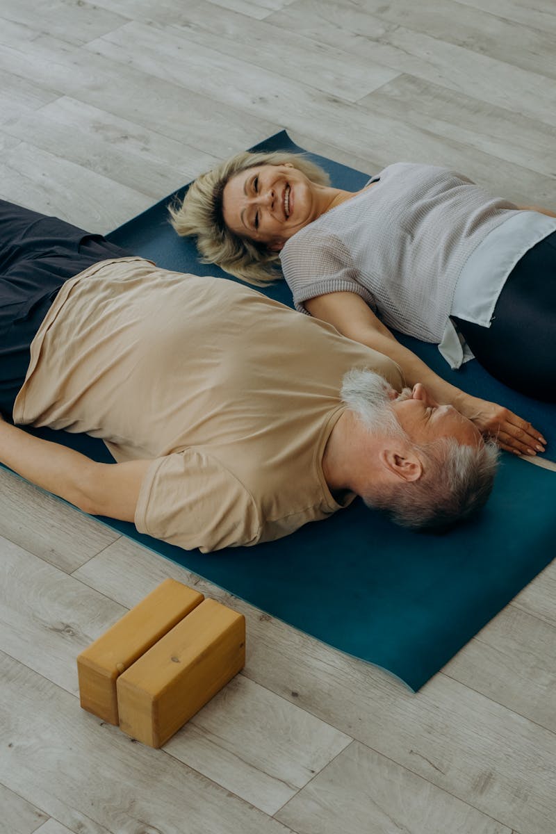 Elderly couple lying on yoga mats, smiling and enjoying relaxation indoors.